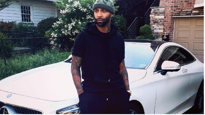 Joe Budden standing in front of his white Mercedez Benz while wearing his black hoddie and trouser